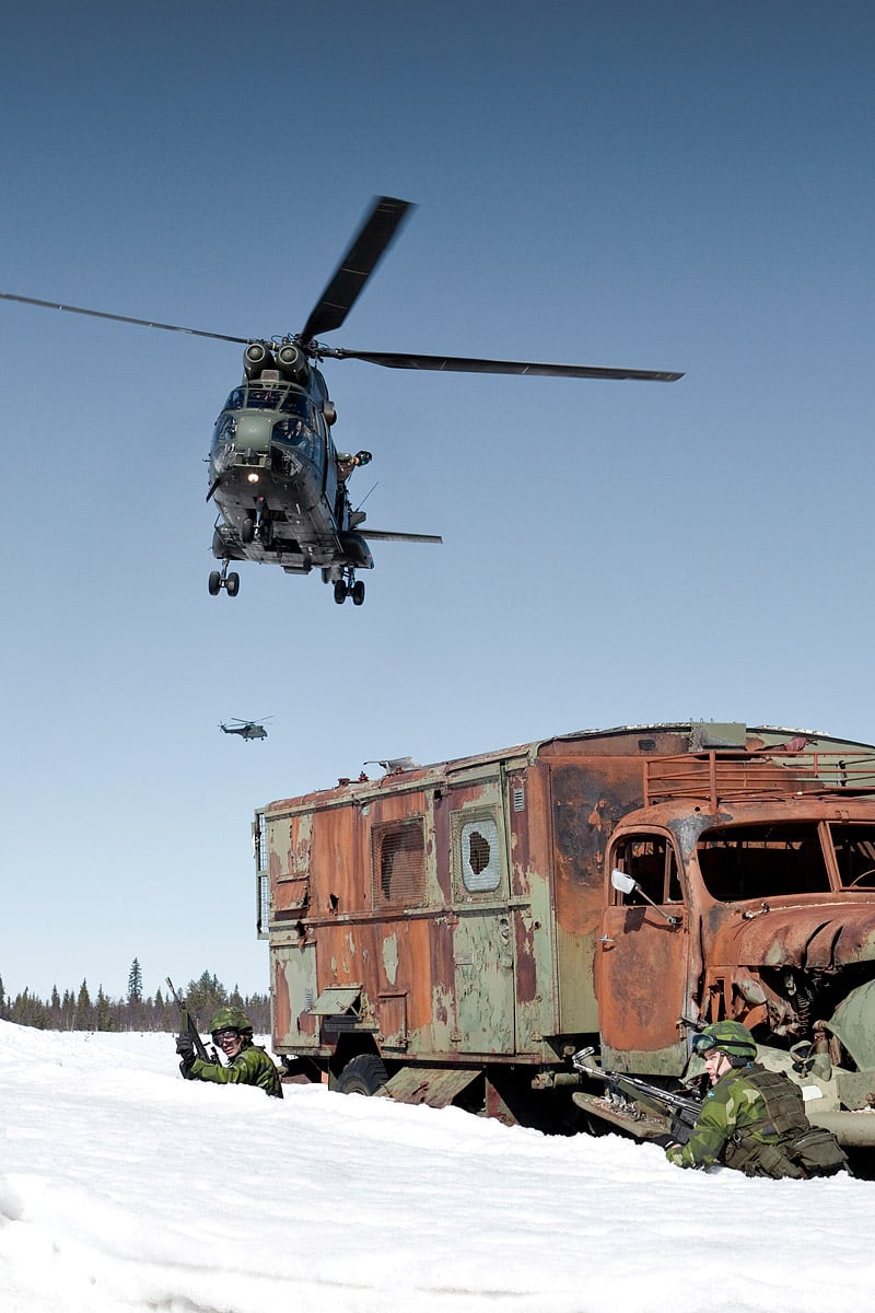 Helicopters in the air over soldiers on the ground, hiding beside a lorry
