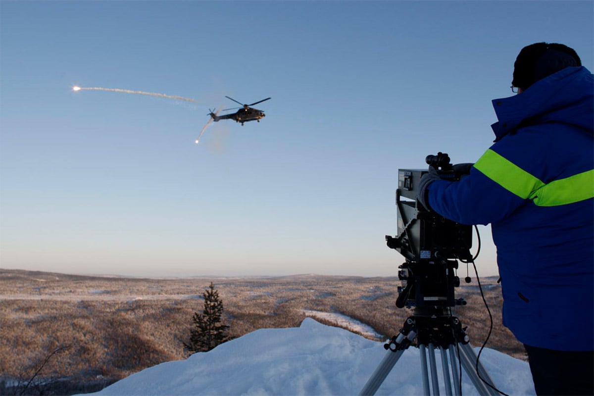 Close-up picture of a Malina operator, with a helicopter ejecting flares in the distance